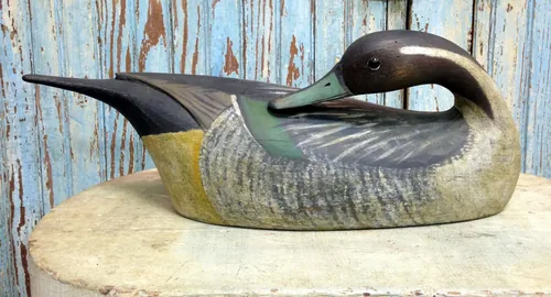 A carved wooden pintail duck decoy with its head turned back preening, worn patina on the paint, on a stone ledge