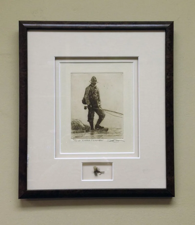 A small sepia etching of a fly fisherman in a dark walnut burl frame with white double mats, and a real hand tied fishing fly mounted in its own window below the etching