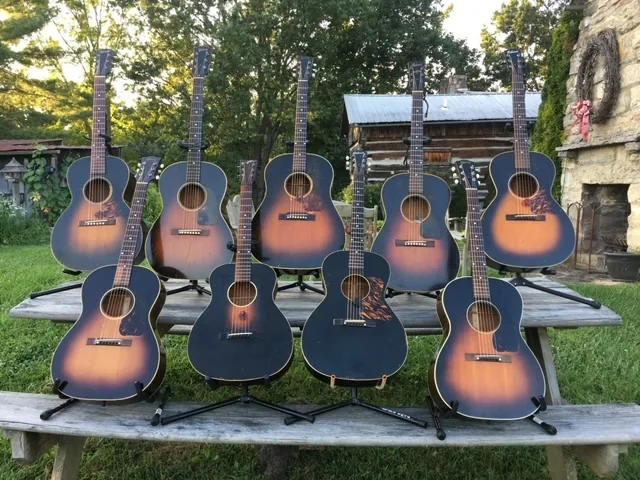 A row of about a dozen vintage acoustic guitars standing on a wooden table outdoors