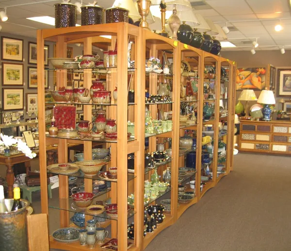 Inside the gallery: tall wooden shelving full of glazed pottery in red, green and blue, framed landscapes on the wall behind, lamps along the top
