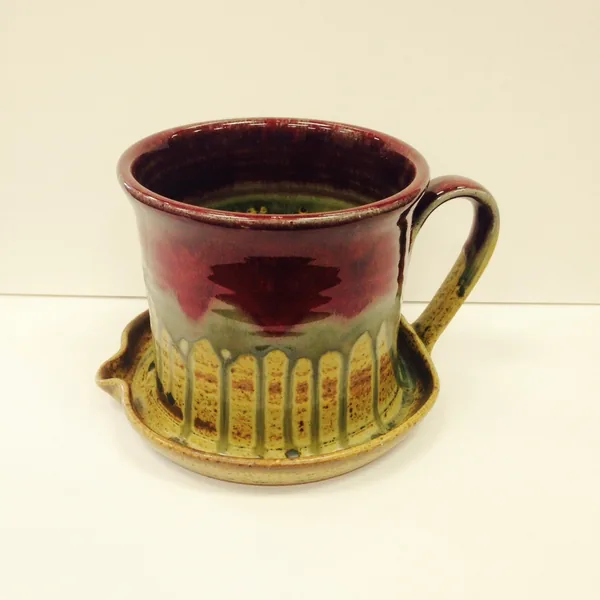 A Ray Pottery bacon cooker with a deep red glaze fading to gold and green, sitting in its saucer
