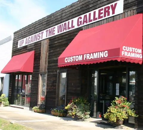The storefront: a dark weathered wood building with a white sign reading Up Against the Wall Gallery and two red awnings reading Custom Framing, flowers in planters by the door